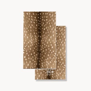 May include: Two rectangular paper napkins featuring a brown and white spotted deer hide pattern. The bottom napkin displays the white text DES ABLENE CO. in a gothic-style font centered near the lower edge of the printed surface.