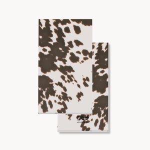 May include: Two rectangular paper napkins featuring a brown and white cow print pattern. The bottom napkin displays the black text DES ARLENE CO. in a stylized gothic font centered near the bottom edge of the cowhide patterned surface.