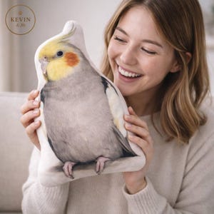 Puede incluir: Un cojín con forma de cacatúa con una impresión detallada del plumaje gris, amarillo y naranja del ave. El cojín está siendo sostenido por una persona.
