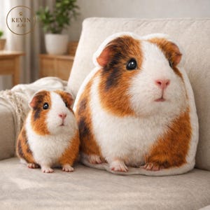Könnte beinhalten: Zwei Meerschweinchen-förmige Plüschkissen. Eines ist kleiner, das andere größer. Die Kissen sind weiß, braun und orange und liegen auf einem beigen Sofa. Die Kissen haben ein realistisches Design. Ein niedliches Wohnaccessoire.