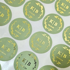 Peut inclure: Plusieurs autocollants ronds vert sauge avec des lettres dorées et une couronne florale. Chaque autocollant porte les initiales "K|B" et la date "05.10.24". Les autocollants sont disposés sur une surface blanche.