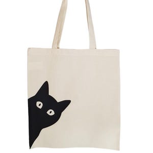 Puede incluir: Una bolsa de lona blanca con un gato negro asom&aacute;ndose por un lado. El gato tiene ojos grandes y redondos.