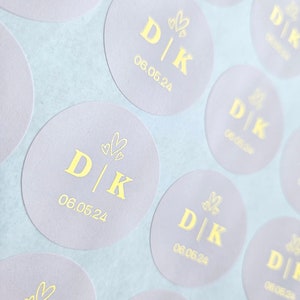 May include: A sheet of round, light grey stickers with gold foil lettering. Each sticker features the initials "D|K" with a small heart design above the letters and the date "06.05.24" below.