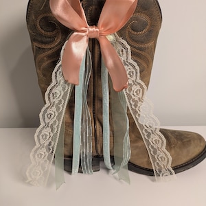 Op de afbeelding: Een bruine leren cowboylaars versierd met een perzikkleurige satijnen strik en delicate kanten en lintaccenten. De laars heeft ingewikkelde stiksels en een klassiek Western-ontwerp, perfect voor evenementen met een landelijk thema.