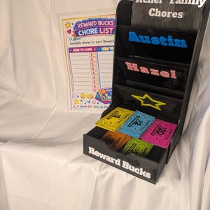 May include: A black wooden chore chart with the text "Keller Family Chores" and the names "Austin" and "Hazel". It includes a "Reward Bucks" box with colorful reward coupons and a chore list.