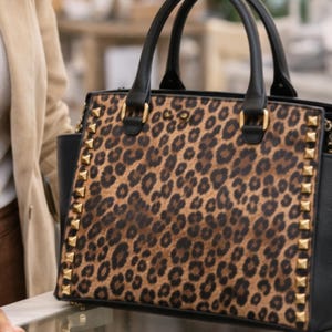 May include: A black and brown leopard print handbag with gold-colored studs and hardware. The bag has black handles and a structured shape. The bag is a stylish accessory.