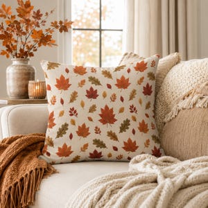 May include: A square throw pillow featuring an off-white background with a repeating pattern of autumn leaves in shades of orange, red, yellow, and olive green. The design includes various leaf shapes, including maple and oak, scattered across the fabric.