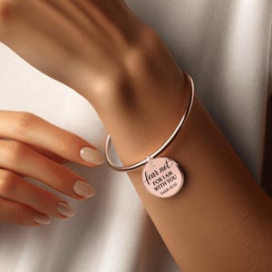 Può includere: Bracciale rigido in oro rosa con un ciondolo circolare. Il ciondolo riporta le parole "fear not FOR I AM WITH YOU Isaiah 41:10" in corsivo nero. Il bracciale è indossato al polso.