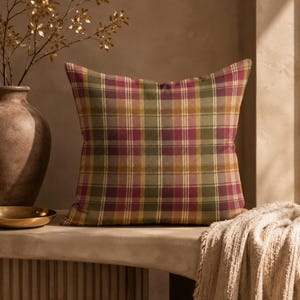 May include: A square scatter cushion featuring a plaid pattern in shades of deep magenta, olive green, and mustard yellow with thin white vertical and horizontal stripes. The fabric has a woven texture and the cushion is filled to a plump shape.