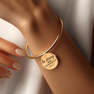 May include: A gold-colored bangle bracelet with a round charm. The charm has the words "be strong AND COURAGEOUS Joshua 1:9" in black script. The bracelet is worn on a wrist.