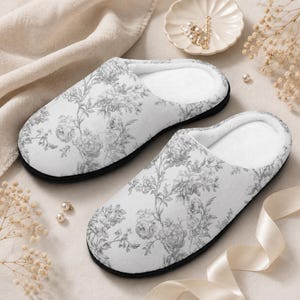 May include: A pair of white floral print slippers with a black sole. The slippers feature a gray rose pattern and a soft, white interior. The slippers are designed for indoor use and comfort.