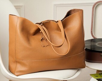 May include: A large, open-top tote bag in a warm tan color, made of textured leather. The bag has two handles and a small leather tag. It sits on a white chair. A vintage record player is in the background.