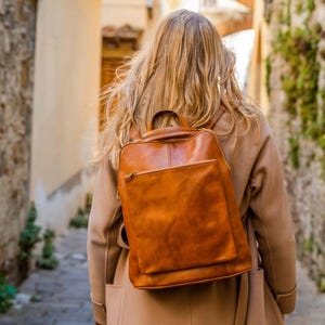 May include: A tan leather backpack featuring a rectangular main compartment, a smaller square front pocket with a zip closure, a top handle, and two shoulder straps. The bag has visible stitching details and a smooth, polished finish.