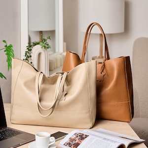 May include: Two leather tote bags in beige and brown tones. The beige bag is in the foreground, with a brown bag behind it. Both bags have handles and a leather tag. A laptop, a cup of coffee, a phone, and a magazine are on the table.