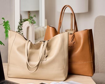 Op de afbeelding: Twee leren tote tassen in beige en bruin. De beige tas staat op de voorgrond, met een bruine tas erachter. Beide tassen hebben handvatten en een leren label. Een laptop, een kopje koffie, een telefoon en een tijdschrift liggen op de tafel.