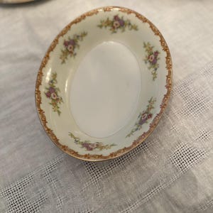 May include: An oval-shaped, off-white porcelain dish with a floral design. The rim features a brown border with a repeating pattern of flowers and foliage in shades of purple, yellow, and green. The dish is part of a set.