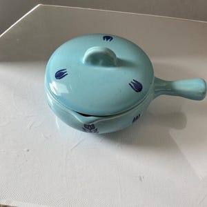Può includere: Una zuppiera in ceramica azzurra con coperchio e manico. La zuppiera e il coperchio sono decorati con disegni di tulipani blu scuro. Il manico è sul lato destro della zuppiera.