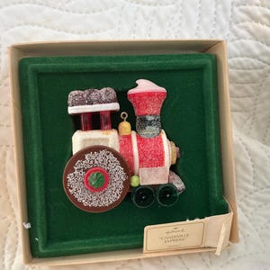 May include: A colorful Christmas ornament in the shape of a train, presented in a box. The train features red, white, and brown elements with a sugary texture. The ornament is set against a green background. The label reads "Candiville Express".