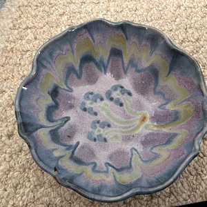 Puede incluir: Un cuenco de cerámica hecho a mano con un diseño floral. El cuenco tiene un borde festoneado y está pintado con tonos de azul, morado y verde. El centro del cuenco tiene un patrón floral con tres pequeñas flores azules.