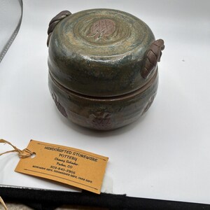 Può includere: Vaso di ceramica in gres fatto a mano con coperchio. Il vaso è rotondo con una glassa verde scuro e marrone. Il coperchio presenta un design strutturato e due manici a forma di corda. L'etichetta recita "HANDCRAFTED STONEWARE POTTERY".