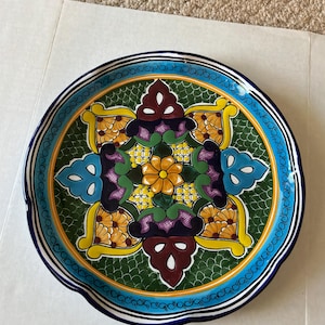 May include: A decorative ceramic plate with a vibrant floral design. The plate features a green center with a symmetrical pattern of yellow, purple, and blue floral elements. A blue border with a white and black trim surrounds the design.
