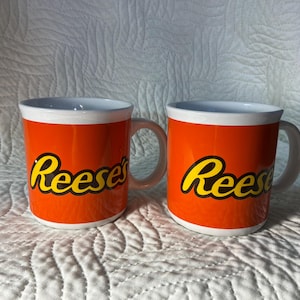 Puede incluir: Dos tazas de cerámica naranja con bordes y asas blancas. Las tazas presentan la palabra "Reese's" en amarillo con un contorno negro. Las tazas están sobre una superficie acolchada blanca.