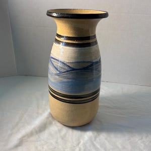 Puede incluir: Un jarrón de cerámica con borde acampanado, que presenta un color base tostado y bandas horizontales de color marrón oscuro alrededor del cuello y el cuerpo. El centro del jarrón muestra un patrón de pinceladas azules que recuerda a colinas.