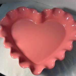 May include: A heart-shaped ceramic dish in a solid light pink color. The dish features a smooth, glossy interior surface and a decorative, ruffled edge that follows the heart contour, creating a scalloped border around the entire perimeter.