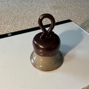 May include: A handcrafted ceramic bell with a dark brown top and a tan base. The bell has a loop handle and a flared bottom. The bell is likely used for decorative purposes.