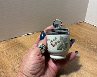 Puede incluir: Un cocedor de huevos de porcelana blanca con una ilustración botánica de moras y hojas verdes. El recipiente tiene una tapa de metal plateado brillante con un asa de anillo circular en la parte superior y una base ensanchada.