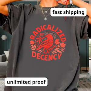 Peut inclure: T-shirt gris foncé avec un motif rouge. Le motif représente un oiseau et les mots "RADICALIZED BY DECENCY" dans un design circulaire. Le t-shirt a des manches courtes et un col rond.