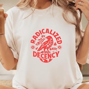 以下が含まれることがあります： 赤いグラフィックの白いTシャツ。グラフィックは、枝をくわえた鳥が、円形のデザインで「RADICALIZED BY BASIC DECENCY」という言葉に囲まれている様子です。このシャツはカジュアルウェアです。
