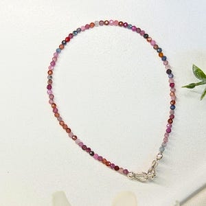 May include: A delicate bracelet composed of small, faceted, multi-coloured gemstone beads in shades of pink, purple, red, and blue. The strand is secured with a polished silver-toned metal lobster claw clasp and a small jump ring connector.