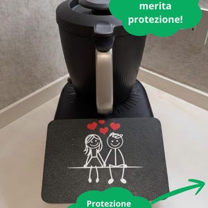 Può includere: Un elettrodomestico da cucina nero con manico argentato e base nera. Un tappetino nero con una coppia di omini stilizzati e cuori è davanti all'elettrodomestico. Il testo in italiano recita: "Il tuo TM7 merita protezione!" e "Massima protezione con i gommini, scoprili!"