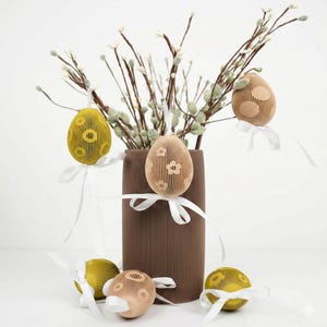 Può includere: Decorazioni pasquali con uova di carta gialle e beige, decorate con nastri bianchi. Le uova presentano motivi ritagliati e sono esposte con rami in un vaso marrone. La composizione è ambientata su uno sfondo bianco.