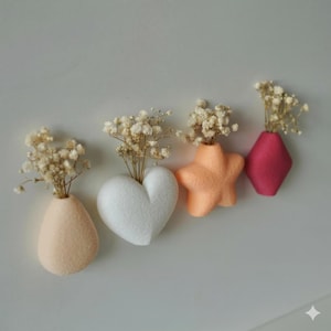 Könnte beinhalten: Vier Filzvasen in Pfirsich, Weiß, Orange und Magenta, jede mit getrockneten weißen Blumen. Die Vasen sind geformt wie ein Tropfen, ein Herz, ein Stern und eine Raute. Die Anordnung befindet sich vor einem schlichten, hellgrauen Hintergrund.