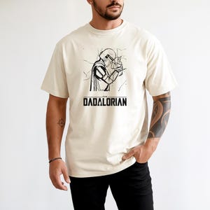 May include: Cream-colored t-shirt featuring a black and white graphic of a helmeted figure holding a small object. The text "THE DADALORIAN" is printed in black below the graphic. The shirt is a relaxed fit.