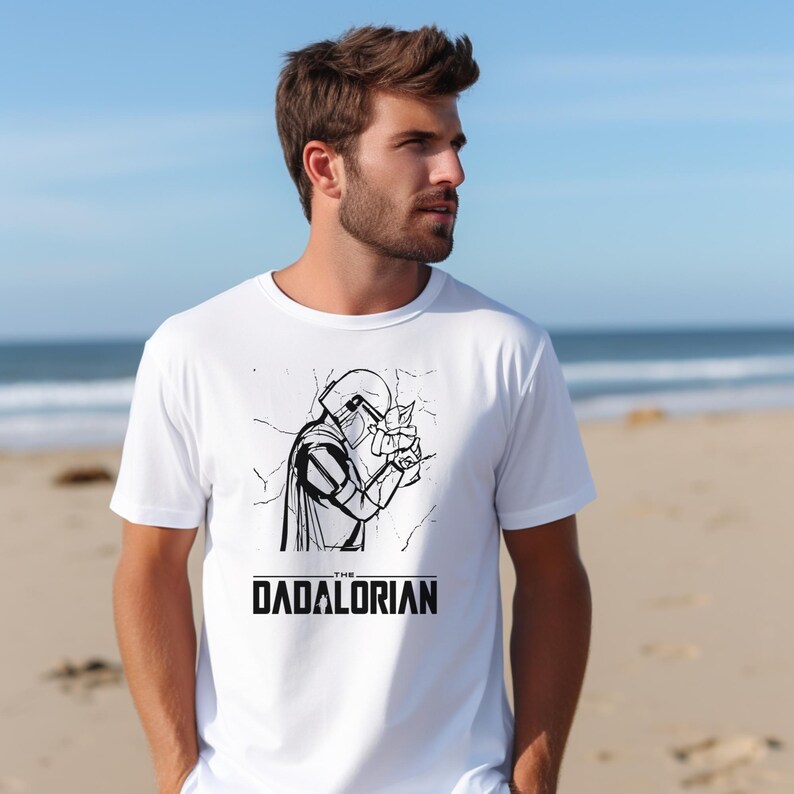 May include: White t-shirt featuring a black and white illustration of a character in a helmet, with the text "THE DADALORIAN" below the graphic. The shirt is short-sleeved and worn by a person on a beach.