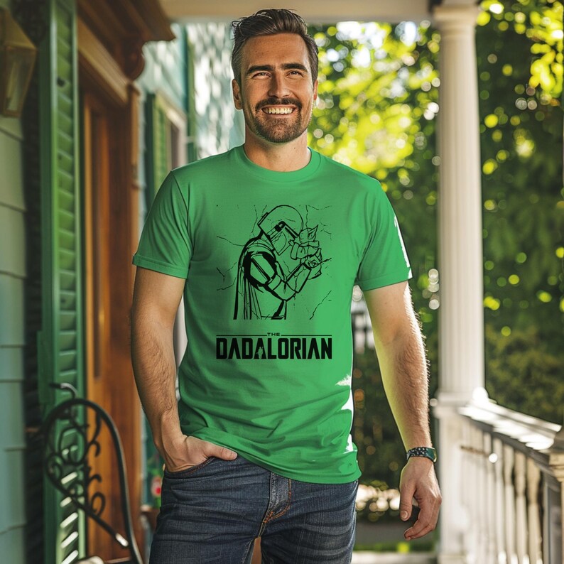 May include: Green t-shirt with a black graphic of a character and the text "The Dadalorian". The shirt is short-sleeved and made of a soft material.