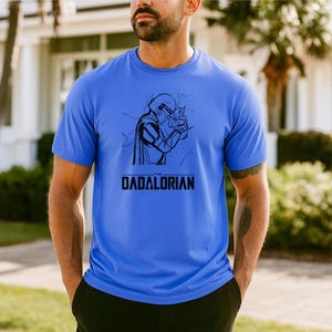 May include: A blue t-shirt featuring a black graphic of a Mandalorian-style character holding a small figure. The text "DADALORIAN" is printed below the graphic in a bold, sans-serif font. The shirt is short-sleeved.
