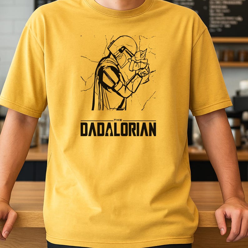 May include: Mustard yellow t-shirt featuring a black graphic of a character holding a small creature, with the text "THE DADALORIAN" below. The design is a parody of a popular science fiction series.