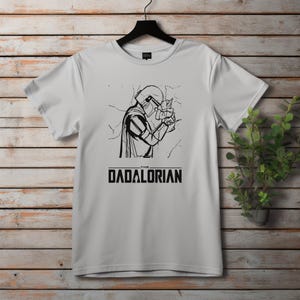May include: White t-shirt featuring a black and white graphic of a character from a science fiction series holding a small creature. The text "THE DADALORIAN" is printed below the graphic in a bold, sans-serif font.