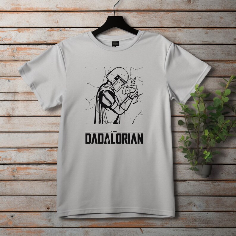 May include: White t-shirt featuring a black and white graphic of a character from a science fiction series holding a small creature. The text "THE DADALORIAN" is printed below the graphic in a bold, sans-serif font.