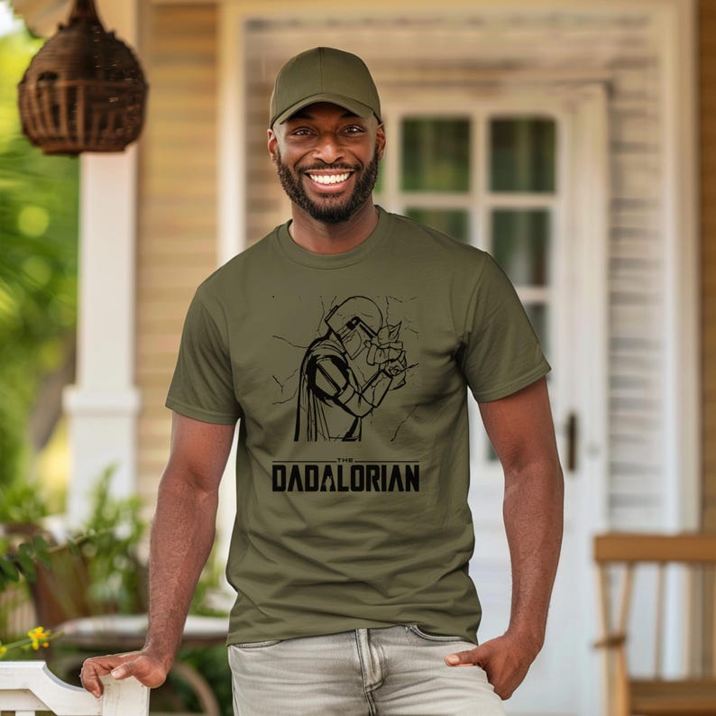 May include: Olive green t-shirt with a black graphic of a helmeted figure and the text "DADALORIAN". The person in the image is wearing a matching olive green baseball cap and gray pants.