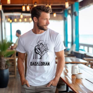 May include: White t-shirt featuring a black and white graphic of a character from a science fiction series, with the text "THE DADALORIAN" below the graphic. The t-shirt is short-sleeved and made of a soft material.