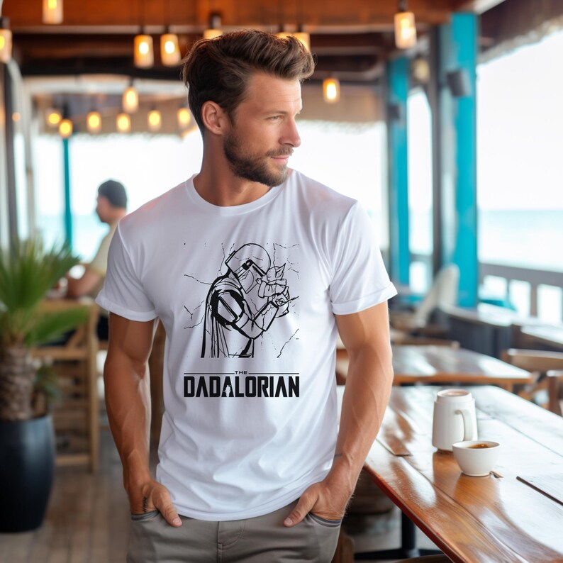 May include: White t-shirt featuring a black and white graphic of a character from a science fiction series, with the text "THE DADALORIAN" below the graphic. The t-shirt is short-sleeved and made of a soft material.