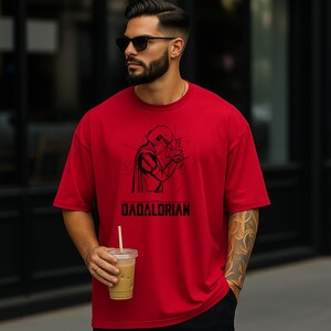 May include: Red t-shirt with a black graphic of a helmeted figure and the word "DADALORIAN" in black block letters. The shirt is a solid red color and has short sleeves.