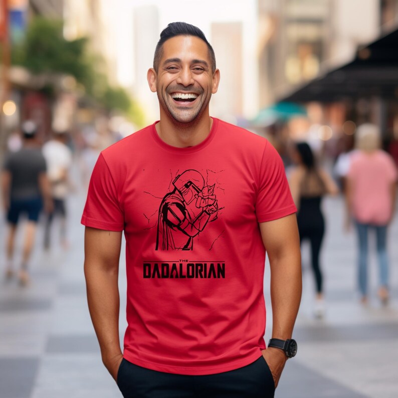 May include: Red t-shirt with a black graphic of a character in a helmet and cape, with the text "The Dadalorian" below. The shirt is short-sleeved and worn by a person smiling in an outdoor setting.