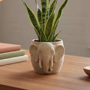 Może przedstawiać: Wężownica z zielono-żółtymi paskami w beżowej doniczce w kształcie słonia. Doniczka stoi na drewnianym biurku obok stosu książek i złotej lampy biurkowej.