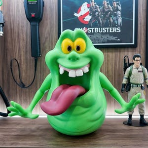 May include: A bright green Slimer character figure with large yellow eyes, a wide mouth showing white teeth, and a long protruding pink tongue. The figure has two outstretched arms with four fingers on each hand and a rounded, blob-like body shape.